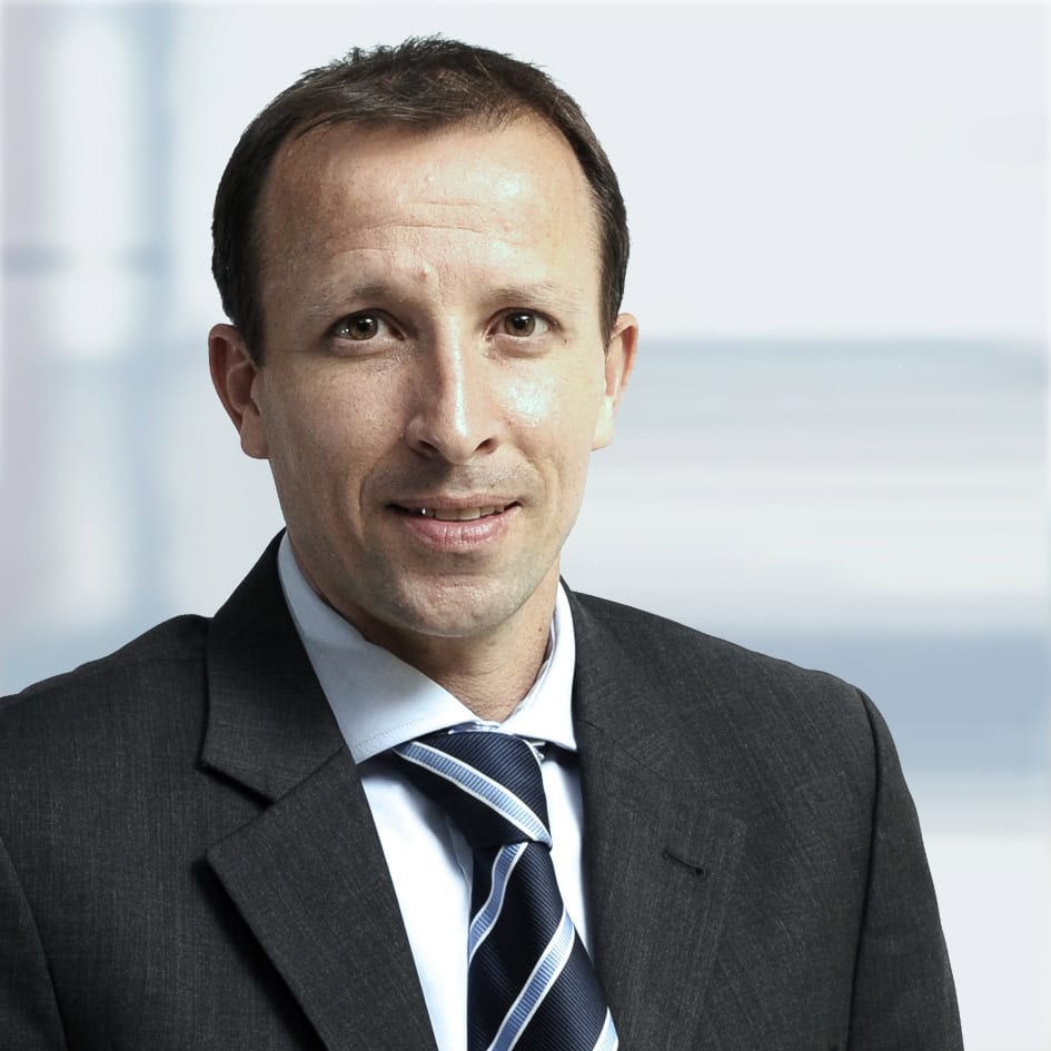 Man wearing a dark suit, light blue shirt, and striped tie, smiling slightly against a blurred background.