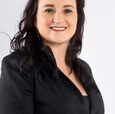 Smiling woman with dark wavy hair wearing a black blazer against a light background.