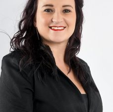 Smiling woman with dark wavy hair wearing a black blazer against a light background.