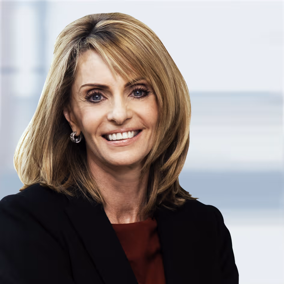 Smiling middle-aged woman with shoulder-length blonde hair, wearing a black blazer and maroon top against a blurred background.