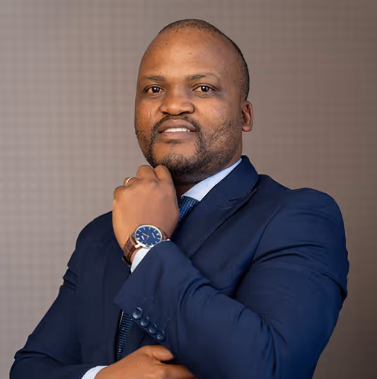 Portrait of a man in a navy blue suit with a watch, posing with his hand near his chin against a neutral background.