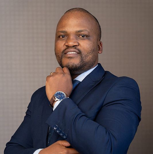 Portrait of a man in a navy blue suit with a watch, posing with his hand near his chin against a neutral background.