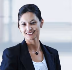 Professional woman with dark hair in a bun, wearing a black blazer and white top, smiling in a bright office setting.