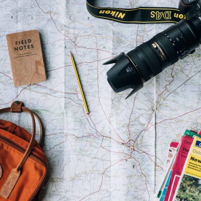 Travel map spread open with a Nikon camera, a pencil, a Field Notes notebook, a brown backpack, and various travel guidebooks.