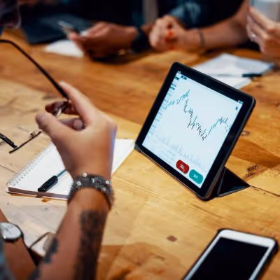 Person reviewing a financial chart on a tablet during a meeting at a wooden table with notebooks and a smartphone nearby.