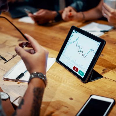 Person reviewing a financial chart on a tablet during a meeting at a wooden table with notebooks and a smartphone nearby.