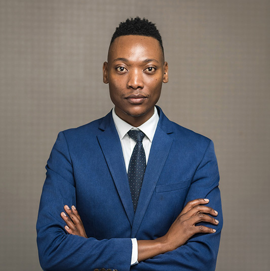 Confident man in a blue suit with a white shirt and black tie standing with arms crossed.