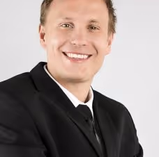 Smiling man wearing a black suit and tie against a plain light background.