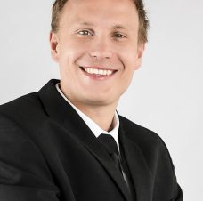 Smiling man wearing a black suit and tie against a plain light background.