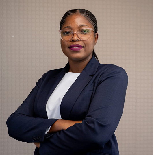 Confident woman with braided hair and glasses wearing a navy blazer and white top, standing with arms crossed.