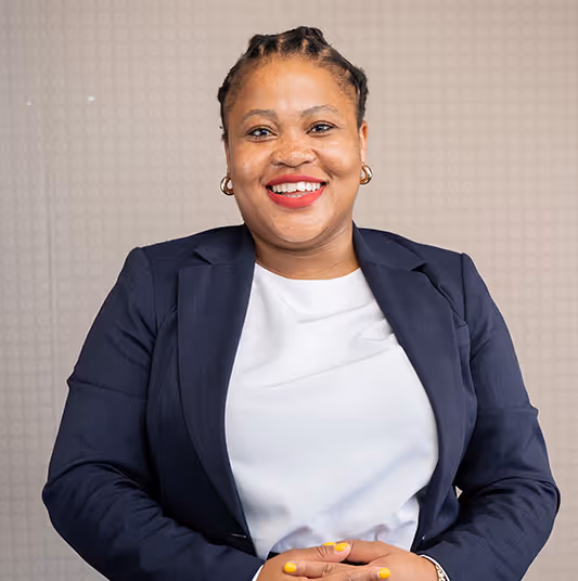 Smiling woman with braided hair, wearing a navy blazer and white top, sitting against a patterned beige background.