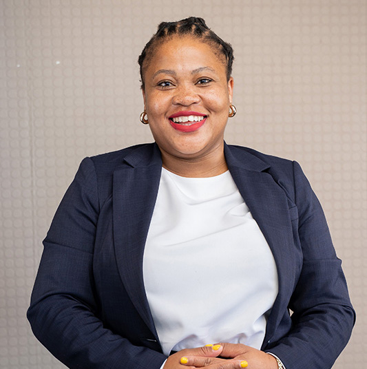 Smiling woman with braided hair, wearing a navy blazer and white top, sitting against a patterned beige background.