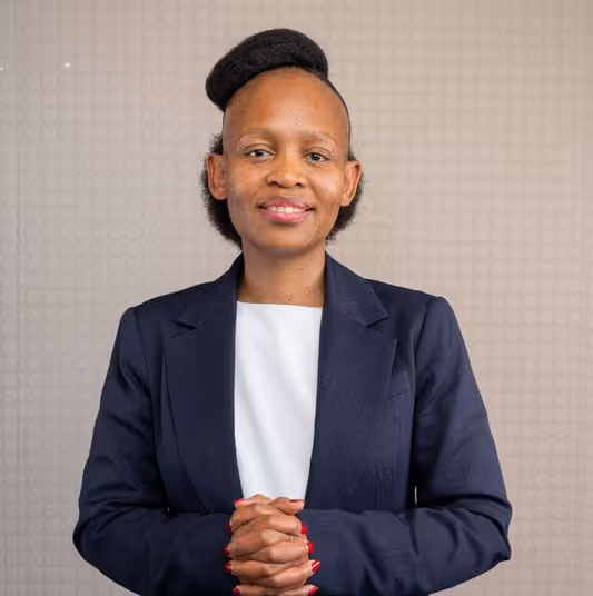 Woman in a navy blazer and white top with hands clasped, smiling against a light patterned background.