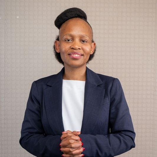 Woman in a navy blazer and white top with hands clasped, smiling against a light patterned background.