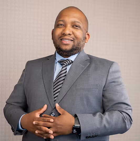 Smiling man in a grey suit jacket, light blue shirt, and striped tie standing with hands clasped in front of a textured beige wall.