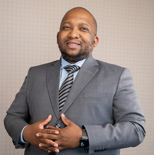 Smiling man in a grey suit jacket, light blue shirt, and striped tie standing with hands clasped in front of a textured beige wall.