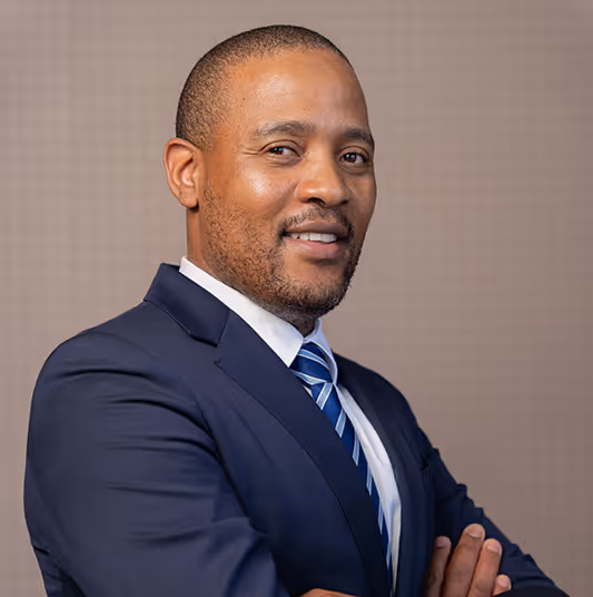 Confident man in a dark suit and striped tie smiling with arms crossed against a neutral background.
