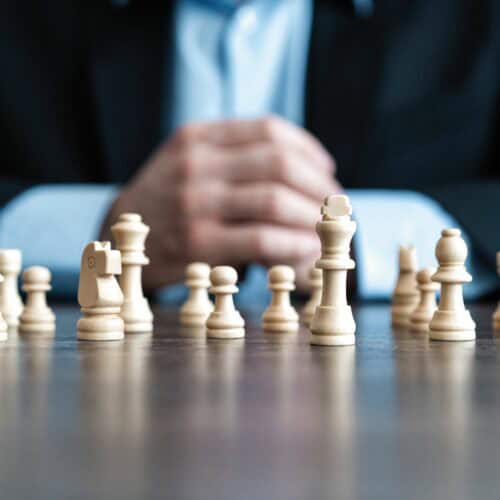 Close-up of wooden chess pieces lined up on a table with a person in a suit blurred in the background.