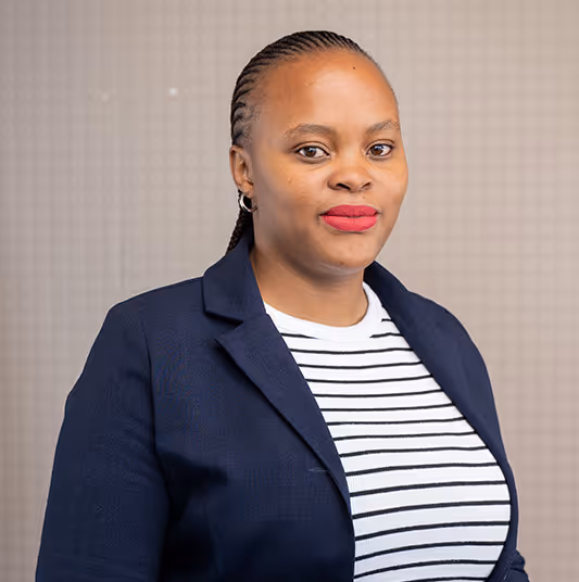 Woman with braided hair wearing a navy blazer and a white striped shirt, standing against a neutral background.
