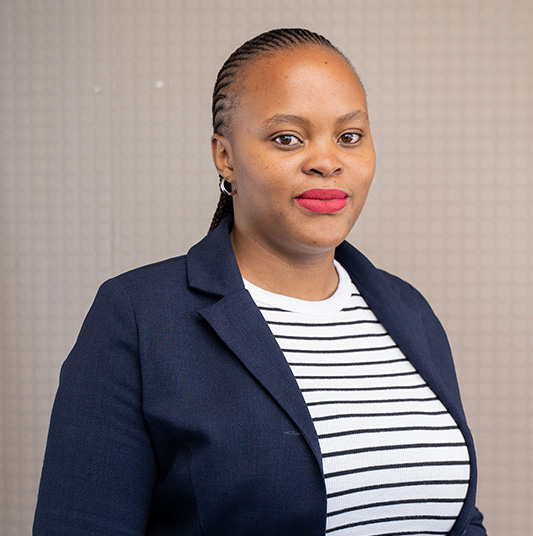 Woman with braided hair wearing a navy blazer and a white striped shirt, standing against a neutral background.