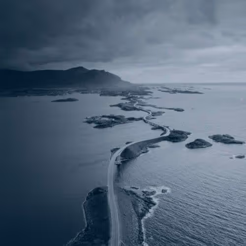 A winding coastal road connecting small islands under a cloudy sky with mountains in the background.