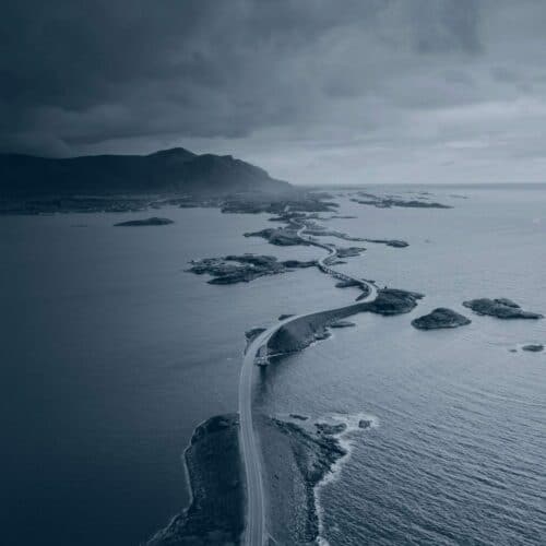 A winding coastal road connecting small islands under a cloudy sky with mountains in the background.