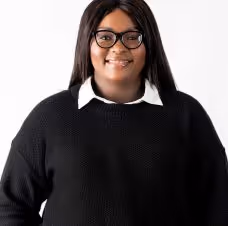 Smiling woman with long dark hair wearing glasses, white collared shirt, and black sweater against a white background.