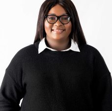 Smiling woman with long dark hair wearing glasses, white collared shirt, and black sweater against a white background.