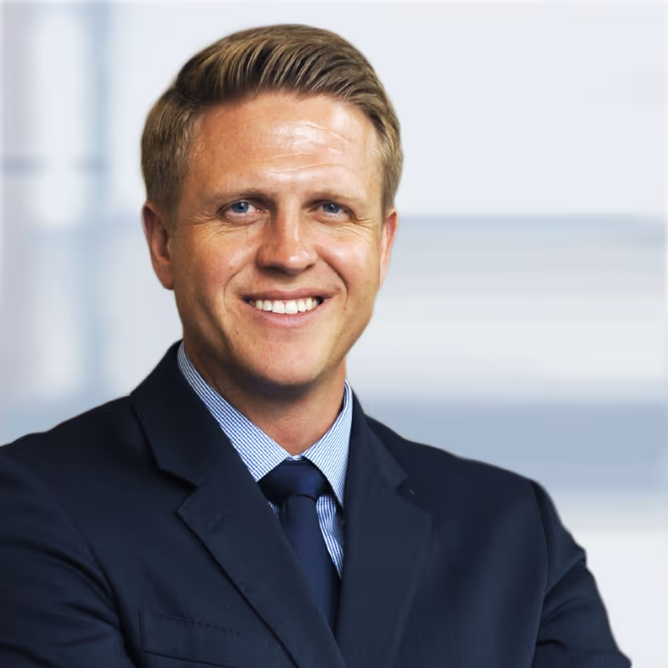 Smiling man with blonde hair wearing a dark suit and blue tie, posing against a blurred light background.