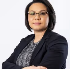 Confident woman with short dark hair wearing glasses, a black blazer, and a patterned top, standing with arms crossed.