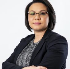 Confident woman with short dark hair wearing glasses, a black blazer, and a patterned top, standing with arms crossed.