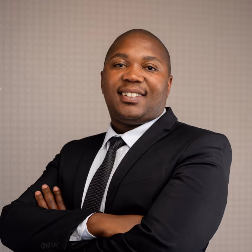 Confident man wearing a black suit and tie, smiling with arms crossed against a neutral patterned background.