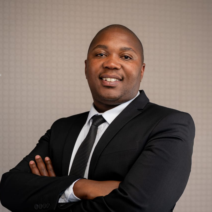 Confident man wearing a black suit and tie, smiling with arms crossed against a neutral patterned background.