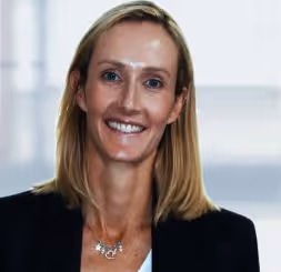 Smiling woman with shoulder-length blonde hair wearing a black blazer and silver necklace against a blurred background.