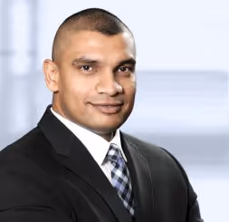 Smiling man in a black suit and checkered tie posing in a bright office setting.