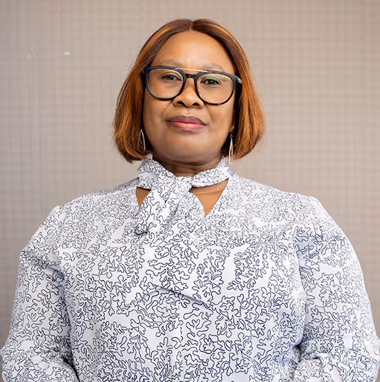 Woman with short brown hair, wearing glasses and a white patterned blouse with a neck tie, posing against a neutral background.
