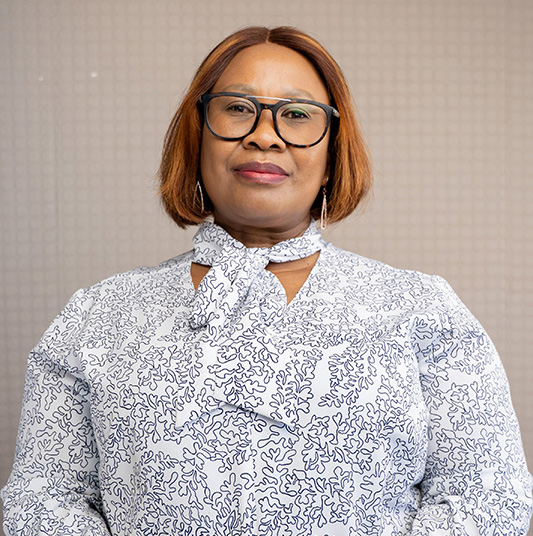 Woman with short brown hair, wearing glasses and a white patterned blouse with a neck tie, posing against a neutral background.