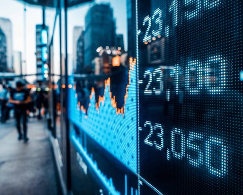 Digital stock market graph and price figures displayed on an urban building glass with blurred city street and pedestrians in background.